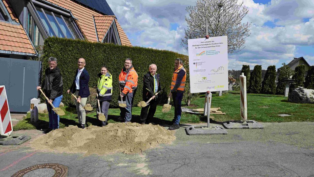 Vertreter der Stadtverwaltung, der Baufirma, des Planungsbüros und des Zweckverbands stehen mit Spaten vor einem Sandhaufen und schaufeln Sand hoch. Rechts am Rand ist ein Förderschild zu sehen. Im Hintergrund steht ein Banner des Planungsbüros.