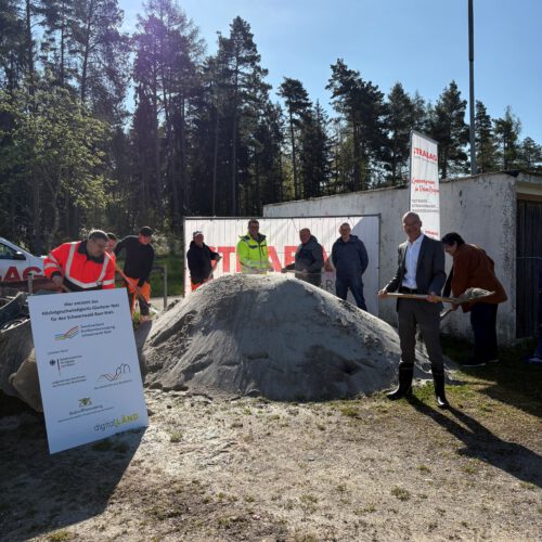 Vertreter der Stadtverwaltung, der Baufirma, des Planungsbüros und des Zweckverbands stehen mit Spaten vor einem Sandhaufen und schaufeln Sand hoch. Links am Rand ist ein Förderschild zu sehen. Im Hintergrund steht ein Banner des Planungsbüros.