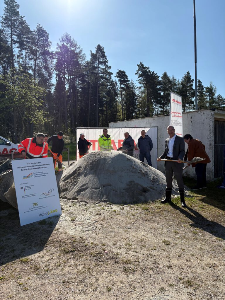Vertreter der Stadtverwaltung, der Baufirma, des Planungsbüros und des Zweckverbands stehen mit Spaten vor einem Sandhaufen und schaufeln Sand hoch. Links am Rand ist ein Förderschild zu sehen. Im Hintergrund steht ein Banner des Planungsbüros.