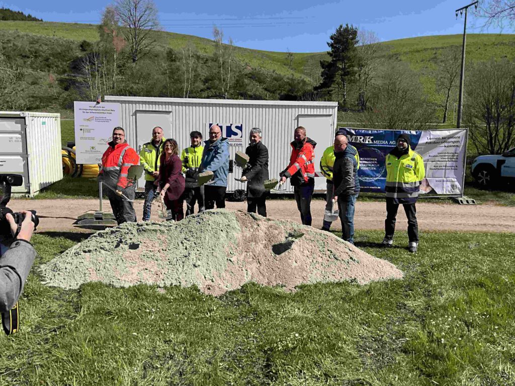 Vertreter der Stadtverwaltung, der Fördergeber, der Baufirma, des Planungsbüros und des Zweckverbands stehen mit Spaten vor einem Sandhaufen und schaufeln Sand hoch. Links am Rand ist ein Förderschild zu sehen.