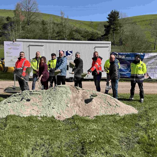 Vertreter der Stadtverwaltung, der Fördergeber, der Baufirma, des Planungsbüros und des Zweckverbands stehen mit Spaten vor einem Sandhaufen und schaufeln Sand hoch. Links am Rand ist ein Förderschild zu sehen.