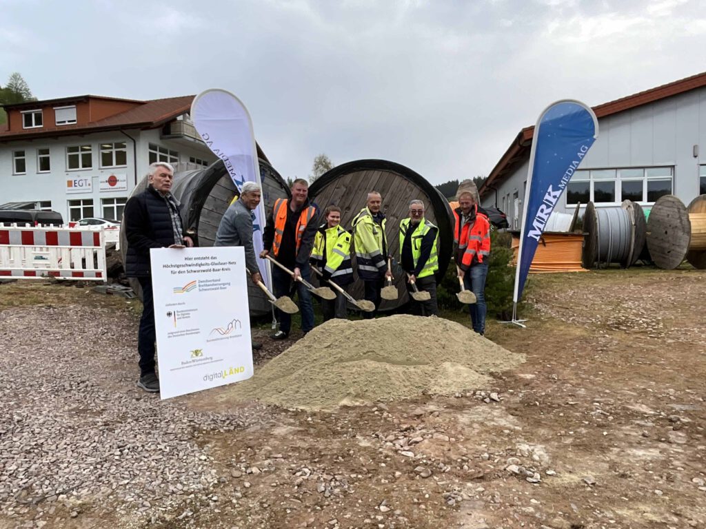 Vertreter der Stadtverwaltung, der Baufirma, des Planungsbüros und des Zweckverbands stehen mit Spaten vor einem Sandhaufen und schaufeln Sand hoch. Links am Rand ist ein Förderschild zu sehen. Im Hintergrund stehen Banner des Planungsbüros.