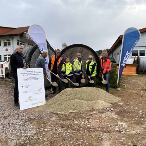 Vertreter der Stadtverwaltung, der Baufirma, des Planungsbüros und des Zweckverbands stehen mit Spaten vor einem Sandhaufen und schaufeln Sand hoch. Links am Rand ist ein Förderschild zu sehen. Im Hintergrund stehen Banner des Planungsbüros.