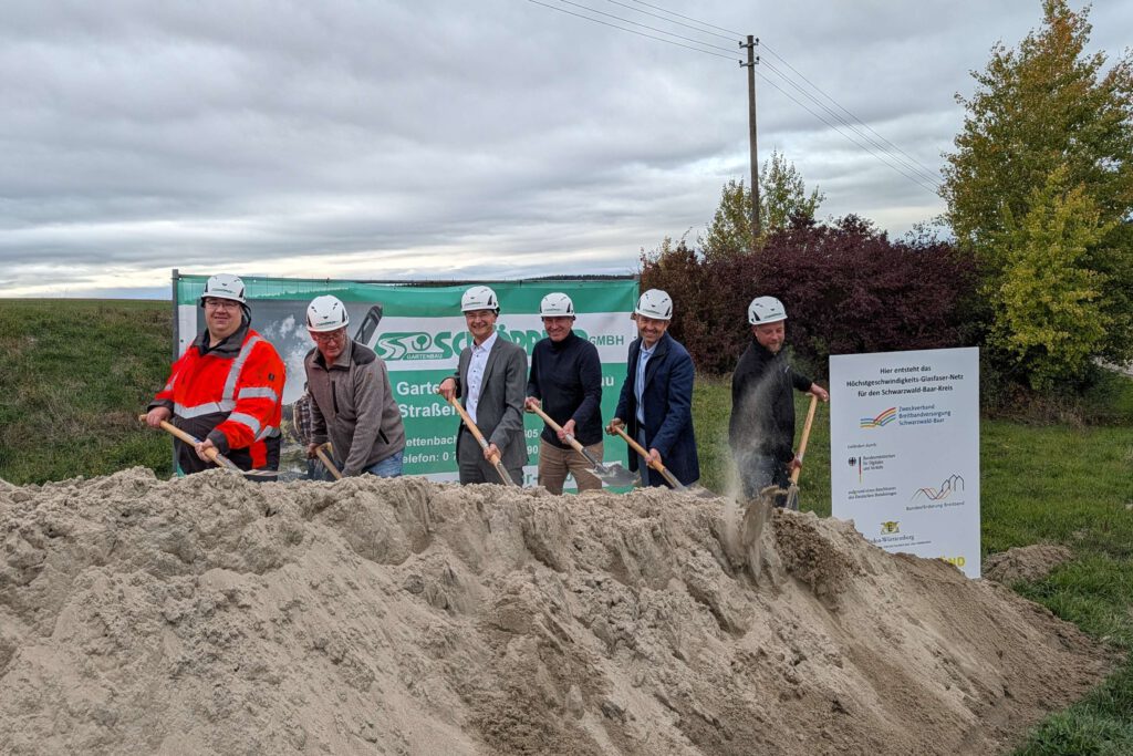 Vertreter der Stadtverwaltung, der Baufirma, des Planungsbüros und des Zweckverbands stehen mit Spaten vor einem Sandhaufen und schaufeln Sand hoch. Rechts am Rand ist ein Förderschild zu sehen. Im Hintergrund steht ein Banner des Planungsbüros.
