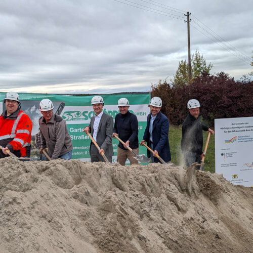 Vertreter der Stadtverwaltung, der Baufirma, des Planungsbüros und des Zweckverbands stehen mit Spaten vor einem Sandhaufen und schaufeln Sand hoch. Rechts am Rand ist ein Förderschild zu sehen. Im Hintergrund steht ein Banner des Planungsbüros.